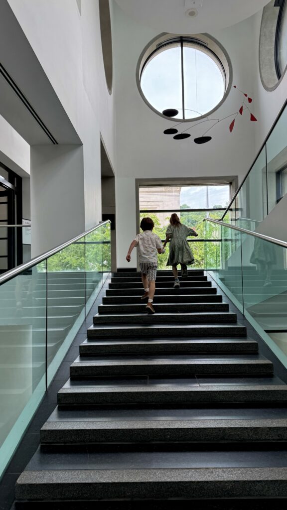 Two children run up a staircase inside the Montreal Museum of Fine Arts, framed by bright natural light, circular windows, and a Calder-style mobile, capturing the energy and curiosity of a long weekend in Montreal.