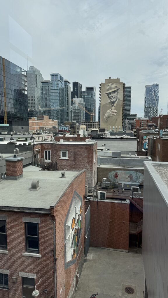 A cityscape view of downtown Montreal featuring a large mural of Leonard Cohen on the side of a high-rise building, seen from the rooftops of older brick buildings with colourful street art, capturing the vibrant contrast of old and new during a long weekend in Montreal. The skyline includes modern glass towers and cranes, hinting at ongoing development.