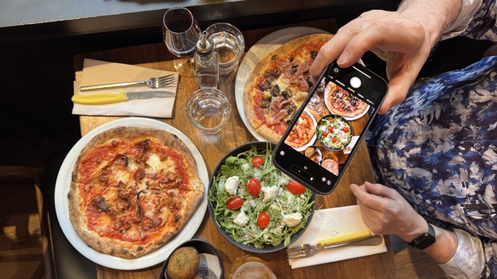 A vibrant table spread at No. 900 Pizza in Montreal featuring two wood-fired pizzas, a fresh arugula salad with cherry tomatoes and mozzarella, and a glass of red wine, as someone snaps a photo to capture the delicious moment during a long weekend in Montreal. The casual, cozy vibe and beautifully plated food make it a perfect foodie stop.