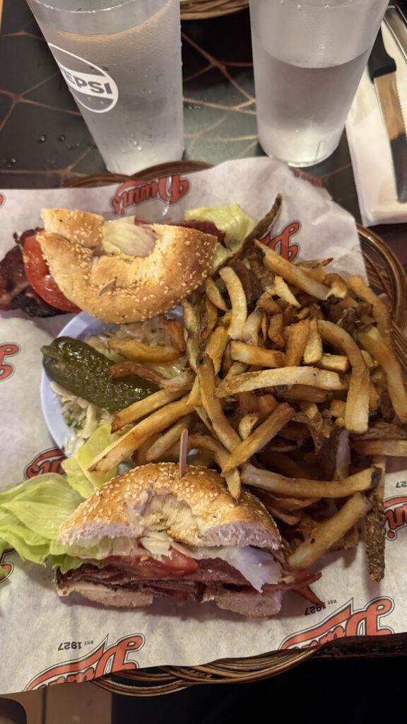 Alt text: A classic deli sandwich on a sesame seed bagel with lettuce, tomato, and sliced meat, served with crispy French fries and a pickle spear at a casual diner. This hearty meal is a great example of popular local fare featured at places to eat in Montreal.