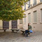 A woman sits on a stone bench in a quiet square in Lyon, with a tree full of green and yellow leaves beside her. She carries a structured blue crossbody bag, a stylish and practical choice that highlights the best travel handbags for Europe.