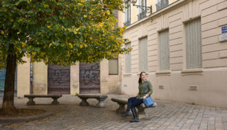 A woman sits on a stone bench in a quiet square in Lyon, with a tree full of green and yellow leaves beside her. She carries a structured blue crossbody bag, a stylish and practical choice that highlights the best travel handbags for Europe.