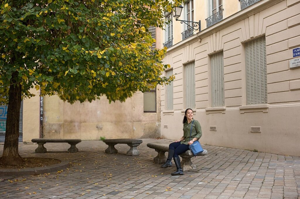 A woman sits on a stone bench in a quiet square in Lyon, with a tree full of green and yellow leaves beside her. She carries a structured blue crossbody bag, a stylish and practical choice that highlights the best travel handbags for Europe.