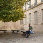A woman sits on a stone bench in a quiet square in Lyon, with a tree full of green and yellow leaves beside her. She carries a structured blue crossbody bag, a stylish and practical choice that highlights the best travel handbags for Europe.