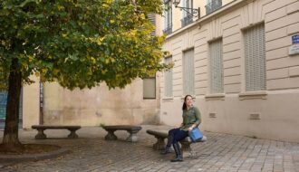 A woman sits on a stone bench in a quiet square in Lyon, with a tree full of green and yellow leaves beside her. She carries a structured blue crossbody bag, a stylish and practical choice that highlights the best travel handbags for Europe.
