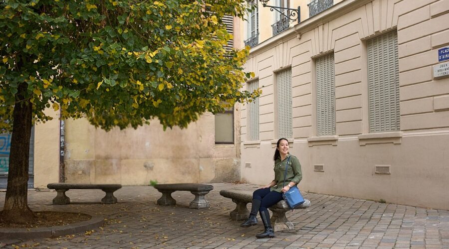 A woman sits on a stone bench in a quiet square in Lyon, with a tree full of green and yellow leaves beside her. She carries a structured blue crossbody bag, a stylish and practical choice that highlights the best travel handbags for Europe.