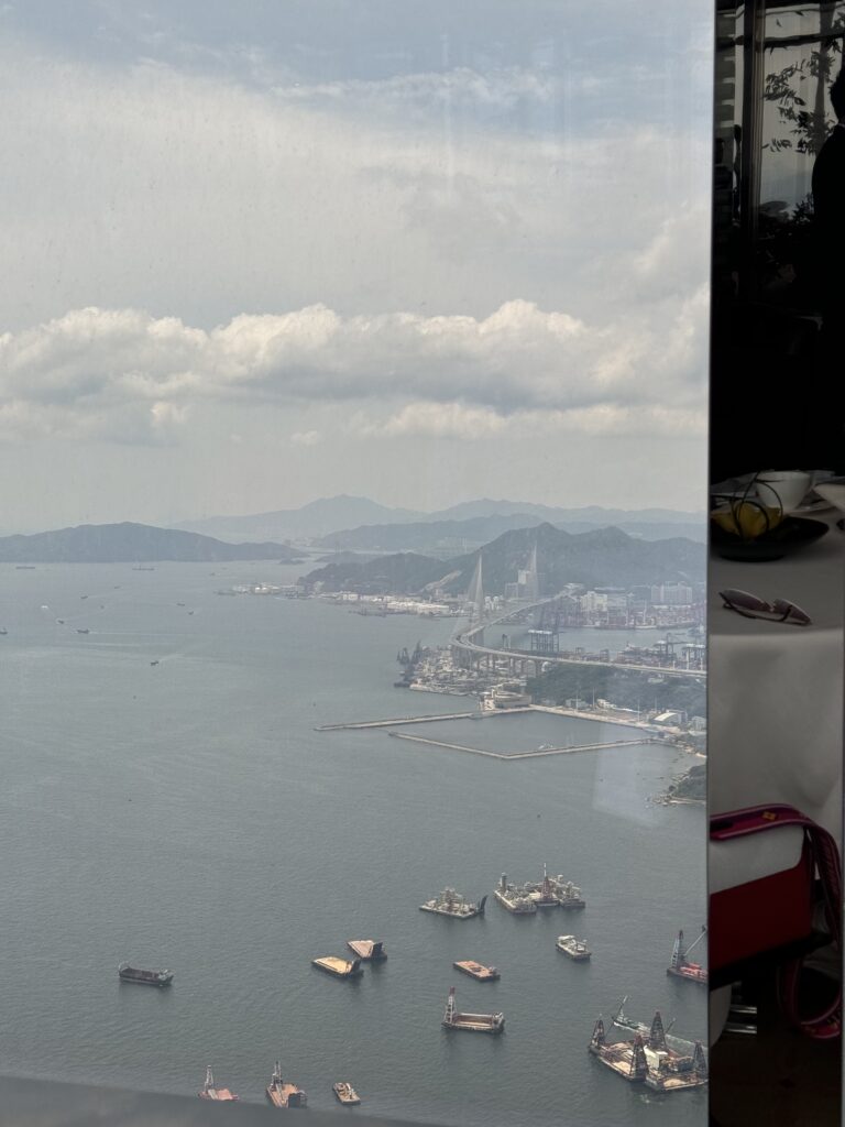 A panoramic view from the Ritz-Carlton Hong Kong shows cargo ships and barges scattered across Victoria Harbour, with the Tsing Ma Bridge stretching toward the mountains in the distance. The scene, framed partly by the restaurant window reflection, offers a striking perspective often recommended in travel tips Hong Kong.