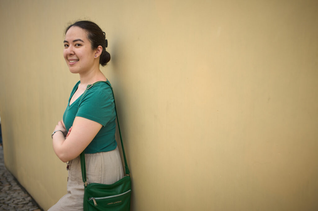 A woman stands against a pale yellow wall smiling, wearing a green top and beige pants in Macau with a crossbody bag. The green leather bag, labeled “Annik Lévesque,” is compact and practical, making it an example of the best travel handbags for Europe.