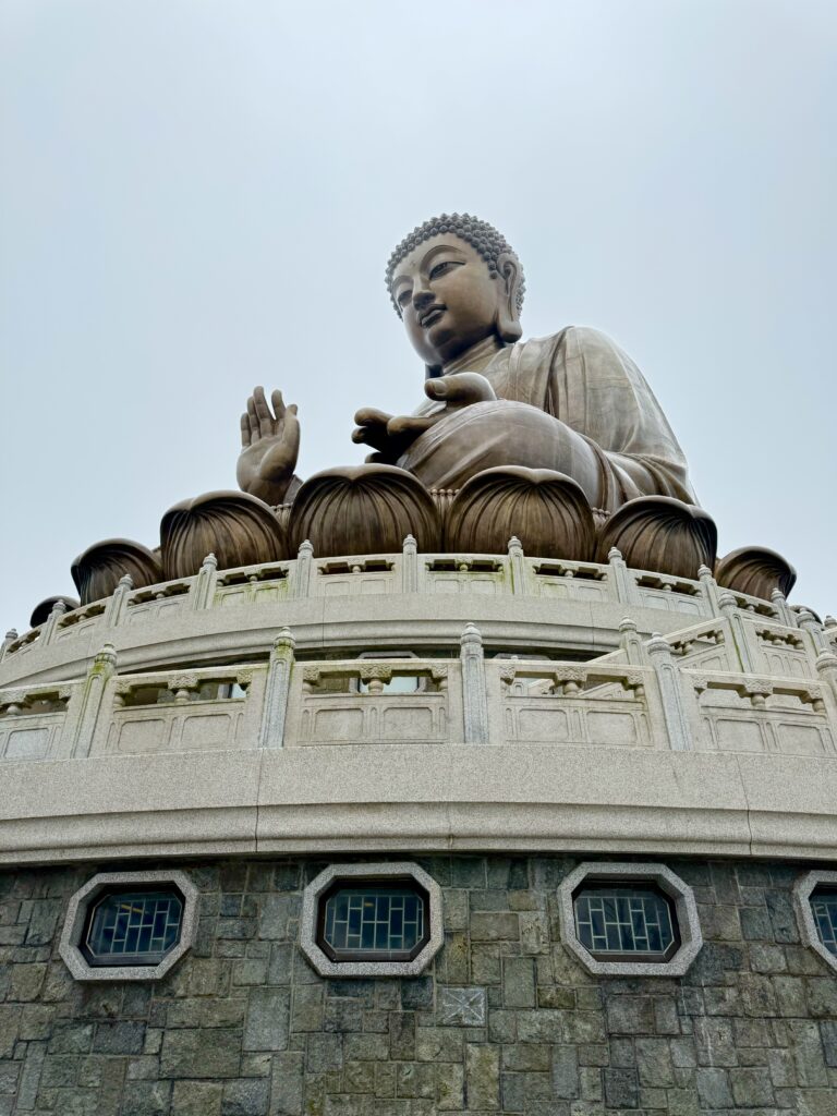 Close-up upward view of the Tian Tan Buddha statue sitting atop a lotus platform with an overcast sky behind. This iconic site on Lantau Island is a must-see during one day in Hong Kong.