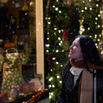 Woman in a winter coat and scarf smiling as snow falls outside a warmly lit storefront decorated with string lights and holiday displays. The cozy evening scene captures the romantic atmosphere of valentine’s day in Canada, blending snowy weather with intimate small town charm.