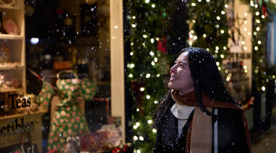 Woman in a winter coat and scarf smiling as snow falls outside a warmly lit storefront decorated with string lights and holiday displays. The cozy evening scene captures the romantic atmosphere of valentine’s day in Canada, blending snowy weather with intimate small town charm.
