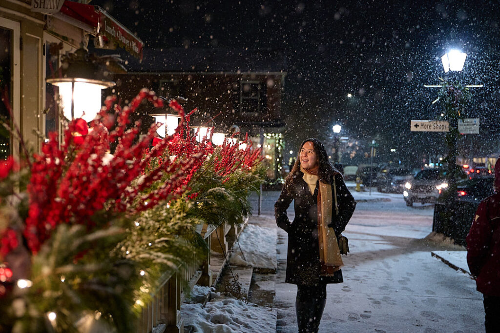 Woman in a dark winter coat and scarf walking along a snowy street in Niagara on the Lake at night as snow falls under glowing streetlights. Festive red planters and warmly lit storefronts create a cozy romantic atmosphere that captures valentine’s day in Canada in this charming heritage town.