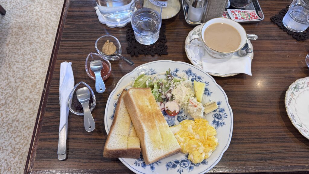 A breakfast set at Bogota Coffee features thick slices of golden toast, fluffy scrambled eggs, and a fresh salad with cabbage, cherry tomatoes, and potato salad on a floral plate. Small dishes of jam and sweet spread sit beside a cup of creamy coffee and a glass of water, capturing the cozy cafe atmosphere that makes Kyoto first time travel feel warm and local.