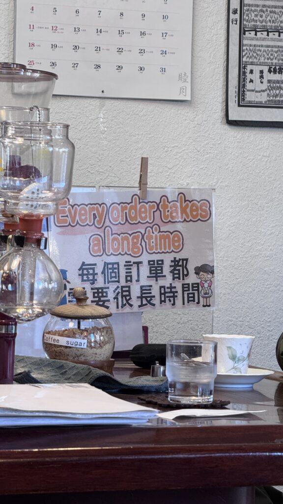 Inside Bogota Coffee, a glass siphon coffee maker sits on a wooden table beside a jar labeled Coffee sugar, a cup and saucer, and a glass of water. A sign reads Every order takes a long time and 每個訂單都要很長時間, highlighting the slow brewed experience that makes Kyoto 1 day itinerary feel authentic and unhurried.