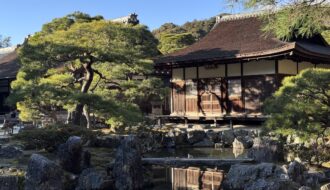 A traditional Kyoto temple building with a sloping tiled roof sits beside a calm pond, surrounded by sculpted pine trees, large natural stones, and a carefully maintained garden. The reflection of the wooden structure in the water captures the peaceful atmosphere that makes Kyoto first time travel so memorable.
