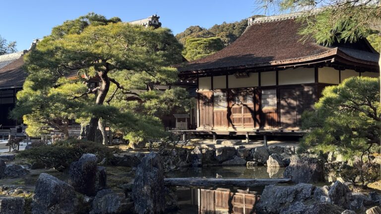 A traditional Kyoto temple building with a sloping tiled roof sits beside a calm pond, surrounded by sculpted pine trees, large natural stones, and a carefully maintained garden. The reflection of the wooden structure in the water captures the peaceful atmosphere that makes Kyoto first time travel so memorable.
