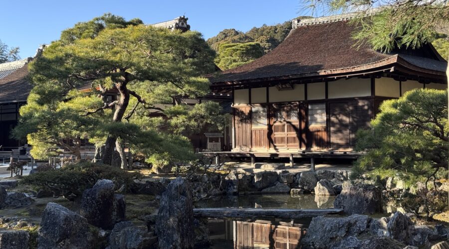 A traditional Kyoto temple building with a sloping tiled roof sits beside a calm pond, surrounded by sculpted pine trees, large natural stones, and a carefully maintained garden. The reflection of the wooden structure in the water captures the peaceful atmosphere that makes Kyoto first time travel so memorable.