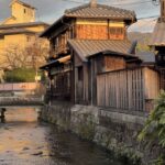 Traditional wooden houses line a narrow canal in Kyoto at sunset, with tiled roofs glowing in warm golden light and a small stone bridge crossing the gently flowing water. This peaceful neighborhood scene captures the timeless charm and quiet beauty you can experience on Kyoto first time travel.