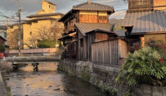 Traditional wooden houses line a narrow canal in Kyoto at sunset, with tiled roofs glowing in warm golden light and a small stone bridge crossing the gently flowing water. This peaceful neighborhood scene captures the timeless charm and quiet beauty you can experience on Kyoto first time travel.