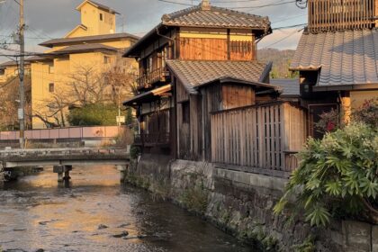 Traditional wooden houses line a narrow canal in Kyoto at sunset, with tiled roofs glowing in warm golden light and a small stone bridge crossing the gently flowing water. This peaceful neighborhood scene captures the timeless charm and quiet beauty you can experience on Kyoto first time travel.