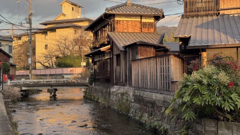 Traditional wooden houses line a narrow canal in Kyoto at sunset, with tiled roofs glowing in warm golden light and a small stone bridge crossing the gently flowing water. This peaceful neighborhood scene captures the timeless charm and quiet beauty you can experience on Kyoto first time travel.