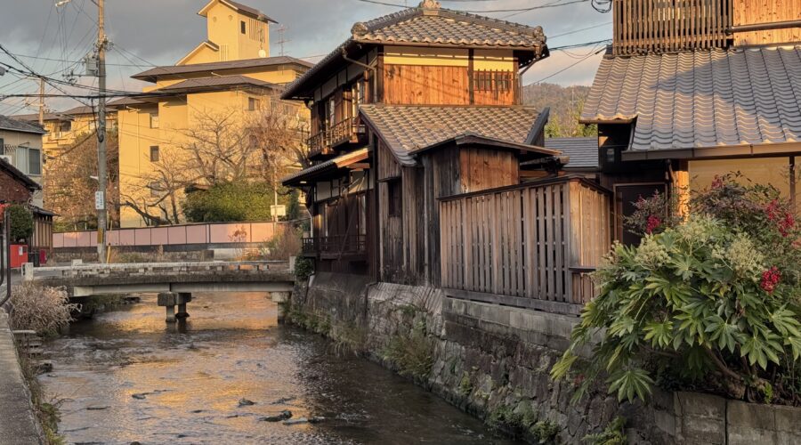 Traditional wooden houses line a narrow canal in Kyoto at sunset, with tiled roofs glowing in warm golden light and a small stone bridge crossing the gently flowing water. This peaceful neighborhood scene captures the timeless charm and quiet beauty you can experience on Kyoto first time travel.
