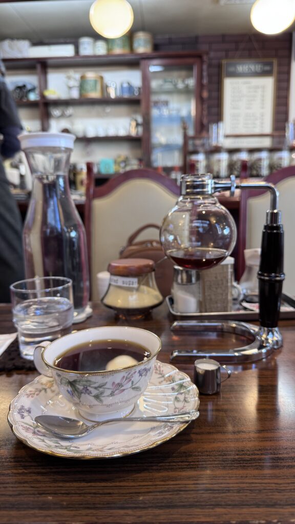 Floral teacup filled with black coffee sits on a wooden table beside a siphon coffee brewer at Coffee Shop Bogota in Kyoto. A calm cafe moment on a Kyoto 3 day itinerary highlighting traditional style coffee preparation.