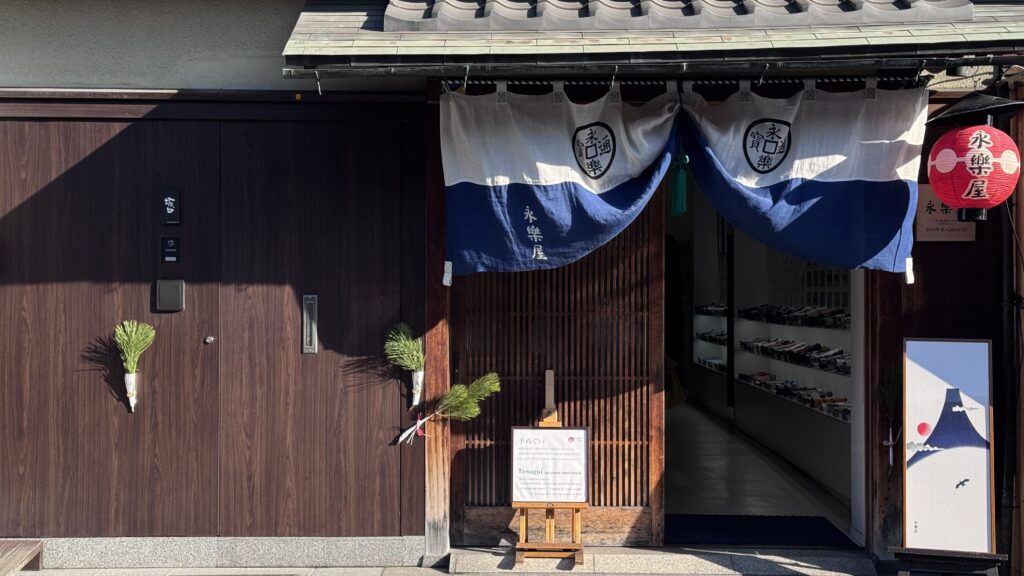 Image: 0c587f583627d8ebc973ca1c715c825e.jpg
Alt text: Traditional wooden storefront in Kyoto with a blue noren curtain displaying the Japanese text “永楽屋” and a red lantern outside the entrance. A signboard reads “Tenugui Japanese hand towel” beside a display of neatly arranged textiles inside, capturing a cultural shopping stop on a Kyoto 3 day itinerary.
