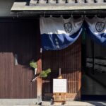 Traditional wooden storefront in Kyoto with a blue noren curtain displaying the Japanese text “永楽屋” and a red lantern outside the entrance. A signboard reads “Tenugui Japanese hand towel” beside a display of neatly arranged textiles inside, capturing a cultural shopping stop on a Kyoto 3 day itinerary.