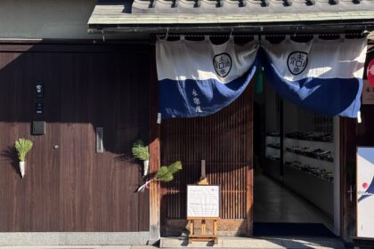 Traditional wooden storefront in Kyoto with a blue noren curtain displaying the Japanese text “永楽屋” and a red lantern outside the entrance. A signboard reads “Tenugui Japanese hand towel” beside a display of neatly arranged textiles inside, capturing a cultural shopping stop on a Kyoto 3 day itinerary.