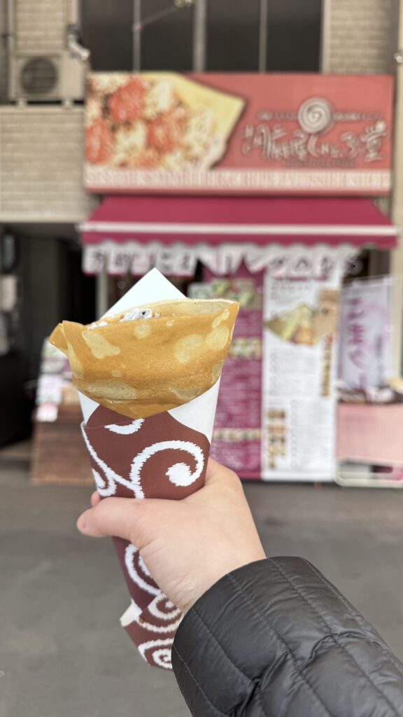Hand holding a freshly made crepe wrapped in paper in front of Gomaya Crepedo storefront with pink signage and menu posters in Kyoto. A quick street food stop on a Kyoto 3 day itinerary highlighting sweet treats and casual eats.