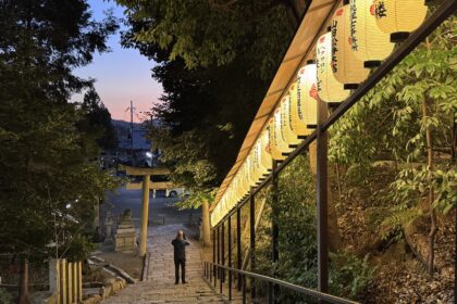Stone steps descend toward a torii gate at Hachi Shrine in Kyoto as rows of glowing paper lanterns illuminate the path at dusk. The peaceful shrine setting showcases a lesser known stop that can be included in a Kyoto 1 day itinerary for travelers exploring the city’s historic neighborhoods after sunset.