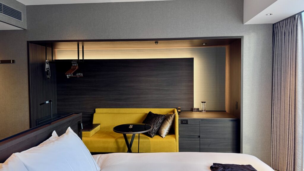 Modern hotel room at Hotel Richmond Premier Shijo in Kyoto featuring a neatly made bed, yellow sofa, and minimalist wood interior design. A comfortable stay option on a Kyoto 3 day itinerary offering a relaxing place to unwind after sightseeing.