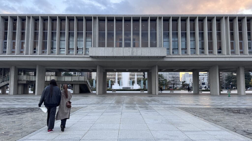 Image: uploaded image
Alt text: Visitors walk toward the Hiroshima Peace Memorial Museum beneath a large modern concrete structure with fountains visible in the distance at sunset. The historic site offers a meaningful cultural experience and is one of the best day trips for first time travellers to Kyoto.