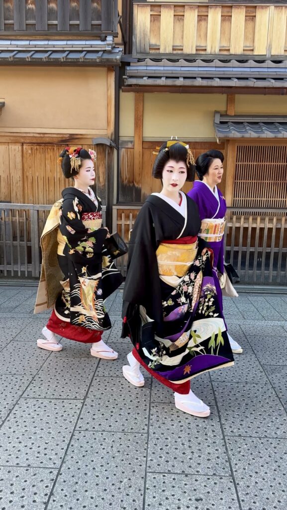 Three geiko and maiko in colourful silk kimonos walk along a stone street in Kyoto, with traditional wooden buildings lining the background. This cultural moment reflects what you might witness on a first time visit to Kyoto in the historic Gion district.