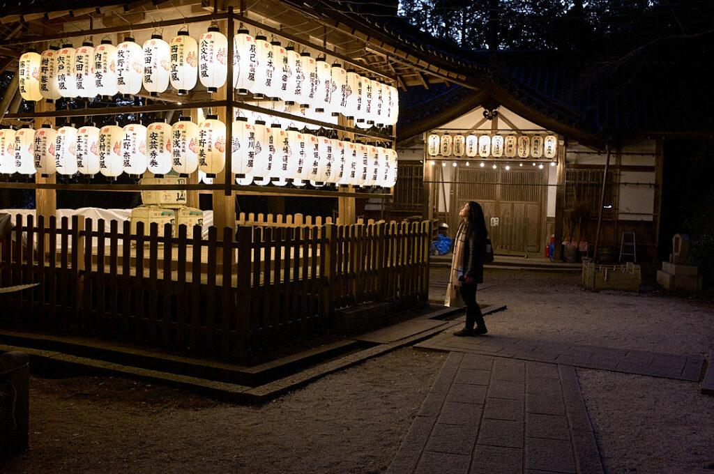 Visitor stands beneath rows of glowing paper lanterns at Hachi Shrine in Kyoto after dark, with traditional shrine buildings softly illuminated in the background. The atmospheric evening scene makes Hachi Shrine a memorable stop on a Kyoto 1 day itinerary, offering a quieter glimpse into the city’s spiritual heritage away from the daytime crowds.