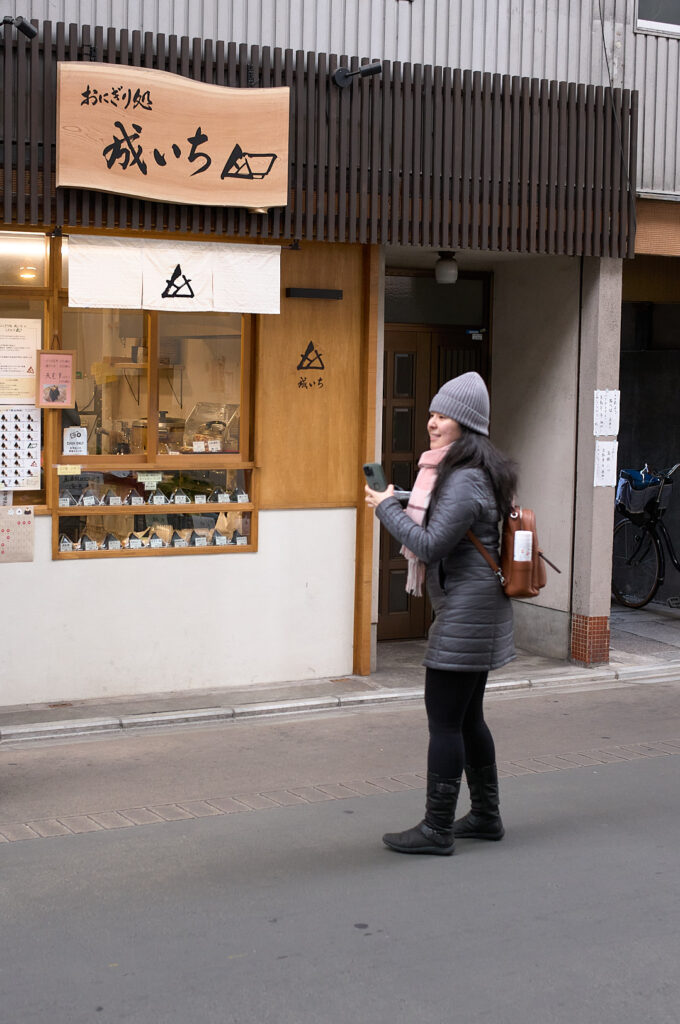 Traveler stands outside a small Kyoto onigiri shop with traditional wooden signage and a display window filled with handmade rice balls. Stopping at local food spots like this adds an authentic culinary experience to a Kyoto 1 day itinerary while exploring the city’s historic streets and neighborhoods.