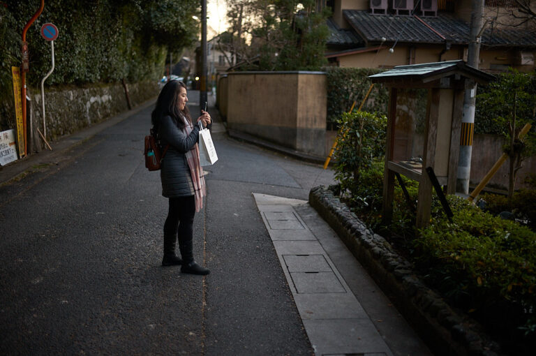 Traveler standing on a quiet residential street in Kyoto at dusk while checking directions on a smartphone. Walking through Kyoto’s historic neighborhoods is one of the best ways of getting around Kyoto, allowing visitors to discover hidden temples, local streets, and cultural landmarks between major attractions.