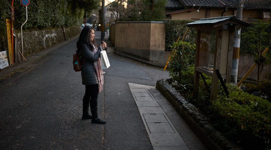 Traveler standing on a quiet residential street in Kyoto at dusk while checking directions on a smartphone. Walking through Kyoto’s historic neighborhoods is one of the best ways of getting around Kyoto, allowing visitors to discover hidden temples, local streets, and cultural landmarks between major attractions.