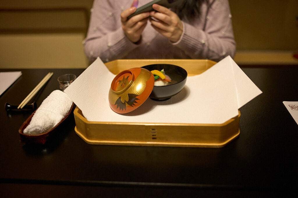 A beautifully presented kaiseki course is served in traditional lacquer bowls on a wooden tray at a Japanese restaurant, while a diner photographs the meal before eating. Experiencing a seasonal kaiseki dinner can be part of a Kyoto budget when chosen as a single splurge meal balanced with lower cost dining throughout the trip.
