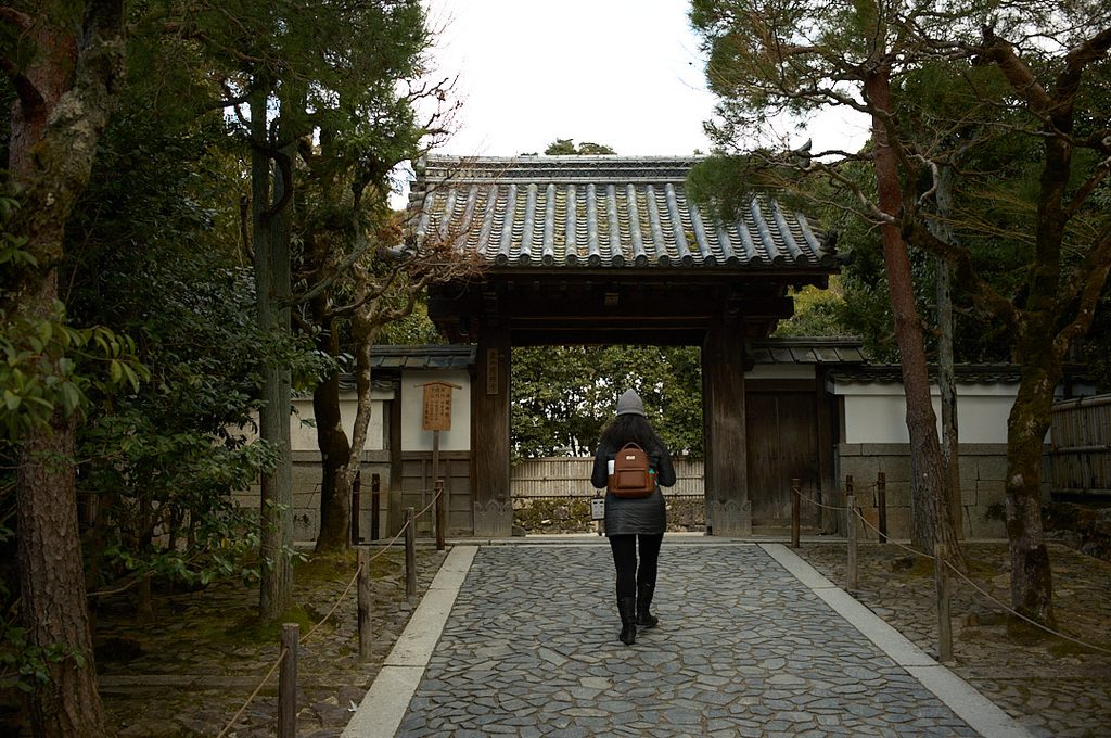 Woman in a winter coat and gray beanie walks along a stone path toward the traditional wooden entrance gate at the Silver Temple in Kyoto. Tall trees and dense greenery surround the tiled roof and temple grounds.