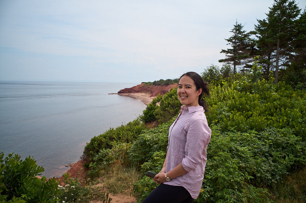 Woman in a light pink shirt smiling on a green coastal cliff overlooking calm water and a red shoreline.